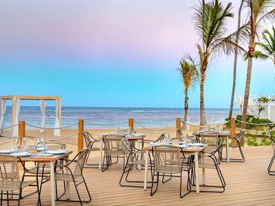 Restaurantterrasse mit Tischen, Stühlen und Palmen am Meer bei klarem Himmel und Sonnenuntergang.