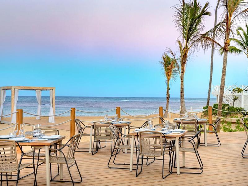 Restaurantterrasse mit Tischen, Stühlen und Palmen am Meer bei klarem Himmel und Sonnenuntergang.