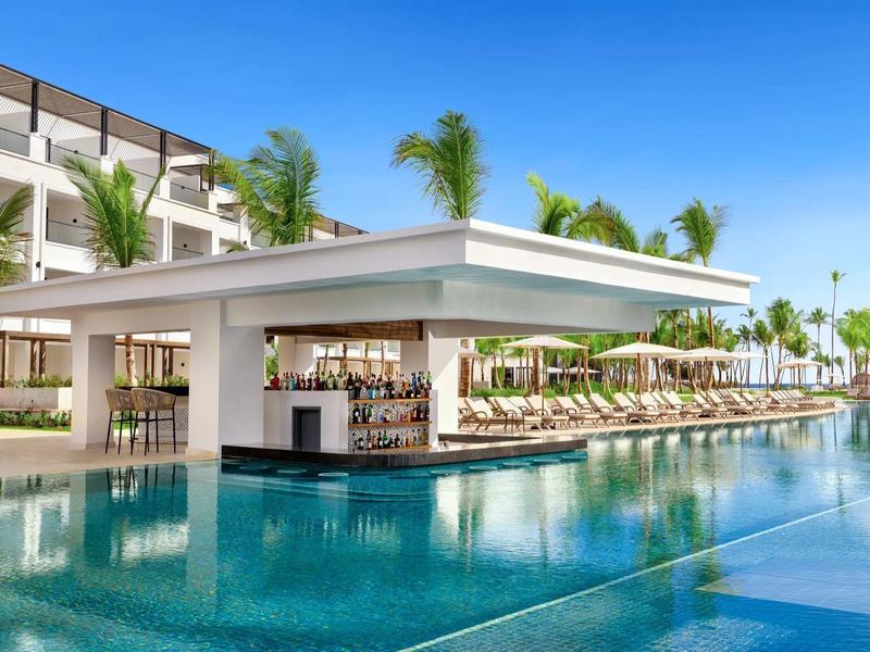 Moderner Hotelpool mit Swim-up-Bar, Liegestühlen und Palmen unter blauem Himmel.