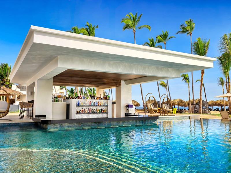 Moderner Pool mit Swim-up-Bar, Sonnenschirmen und Palmen unter klarem, blauem Himmel.