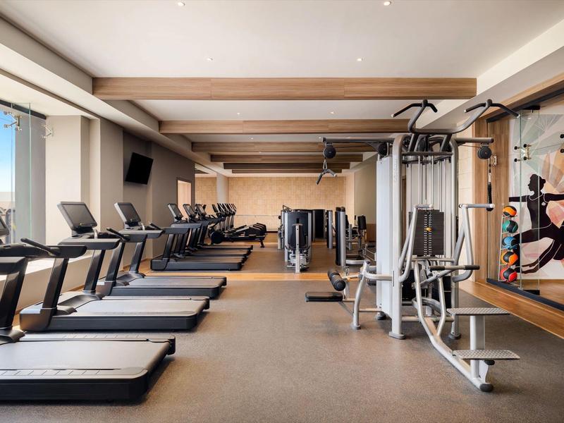 Moderner Fitnessraum mit Laufbändern, Kraftgeräten und großen Fenstern für Tageslicht.