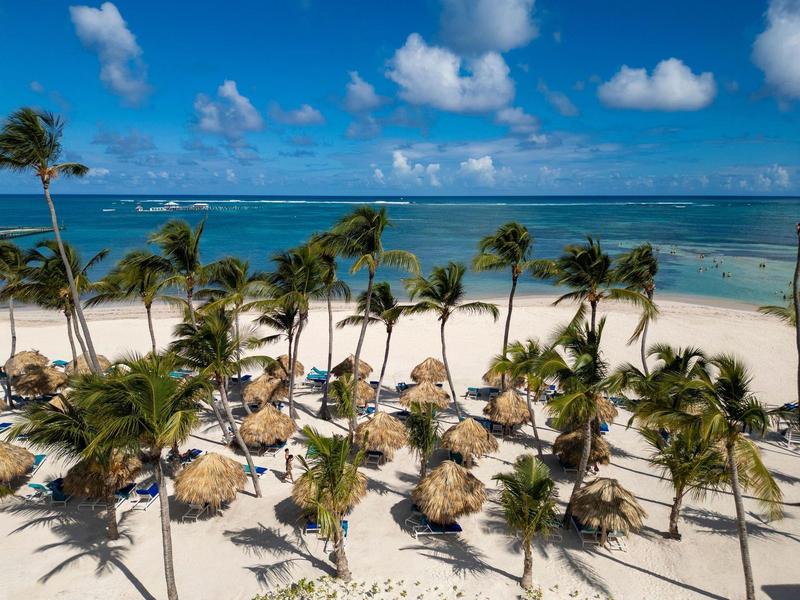 Strand mit vielen Palmen, kleinen Strohhütten, weißem Sand und blauem Himmel mit Wolken.