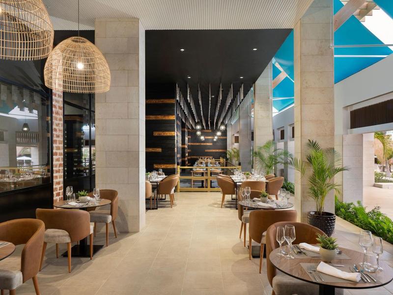 Modernes Restaurant mit Holz- und Korbmöbeln, hellem Boden und großem Fenster für Tageslicht.