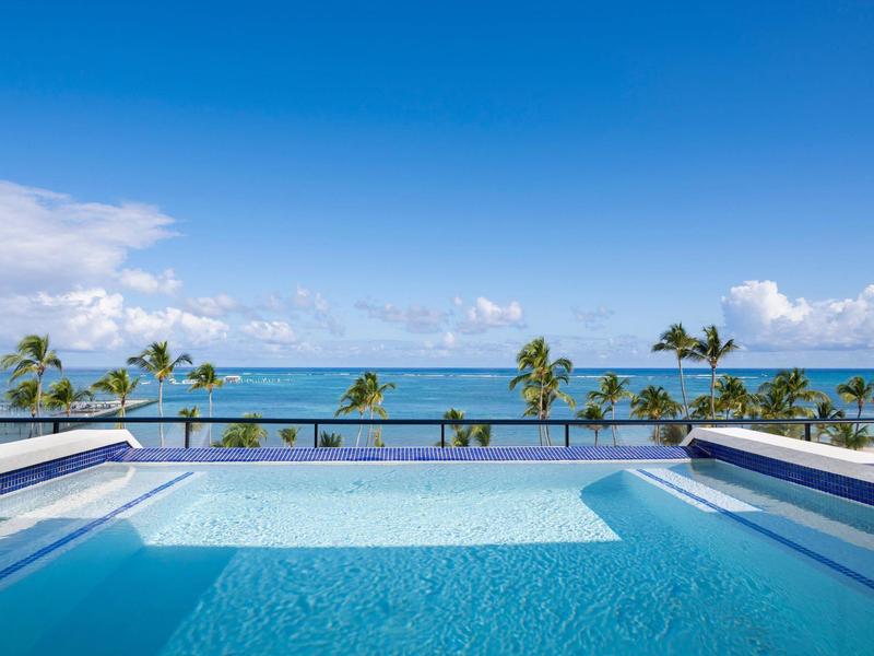 Schwimmbad mit klar blauem Wasser, Blick auf Palmen und den weiten Ozean unter blauem Himmel.