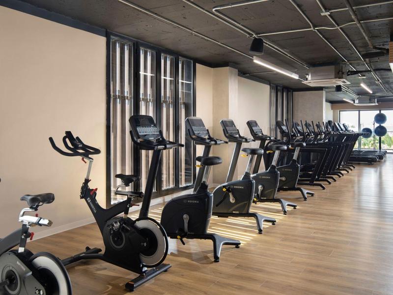 Großer heller Fitnessraum mit Holzfußboden und langen Fensterreihen, voll mit schwarzen Trainingsgeräten.