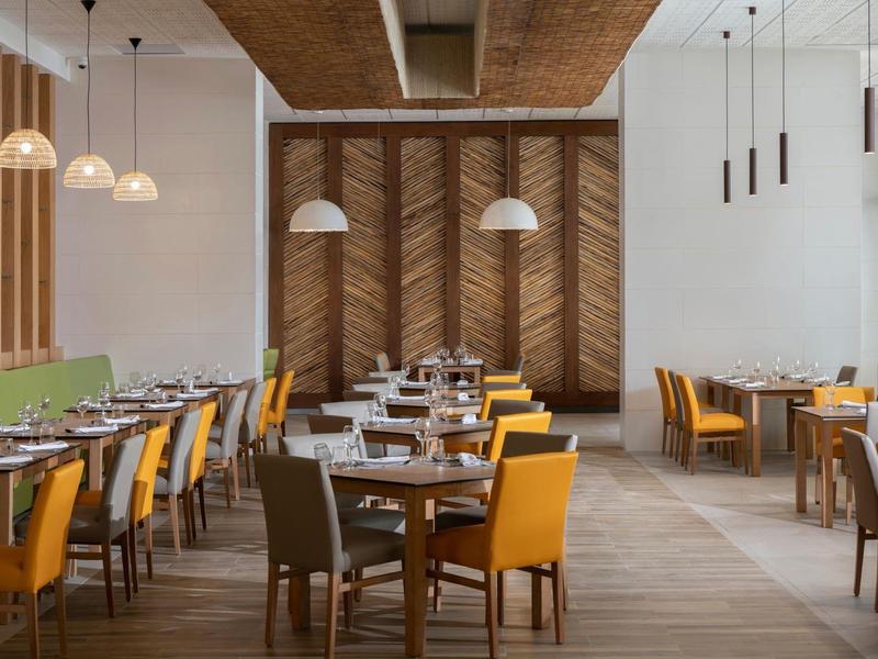 Modernes Restaurant mit Holztischen, bunten Stühlen und hängenden Lampen in hellem Raum.