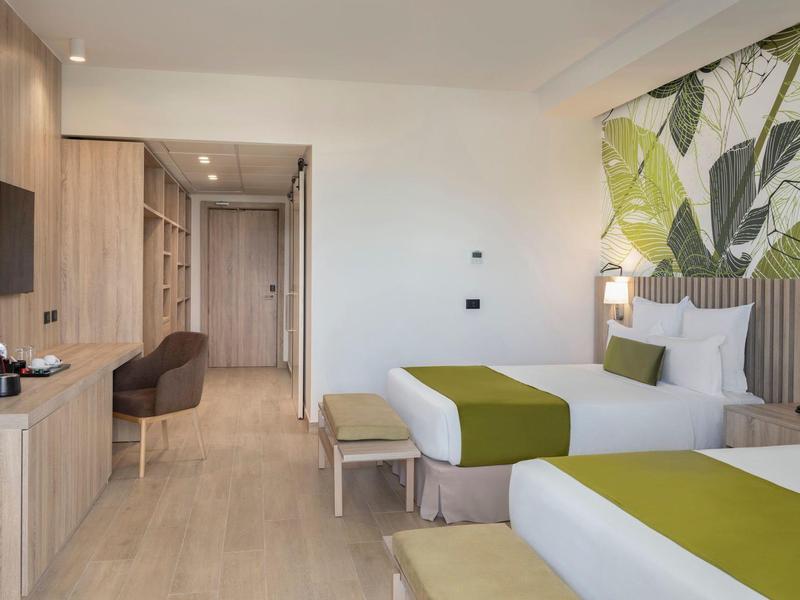 Helle Hotelzimmer mit zwei Einzelbetten, grünen Akzenten, Schreibtisch und großem Pflanzenbild an der Wand.