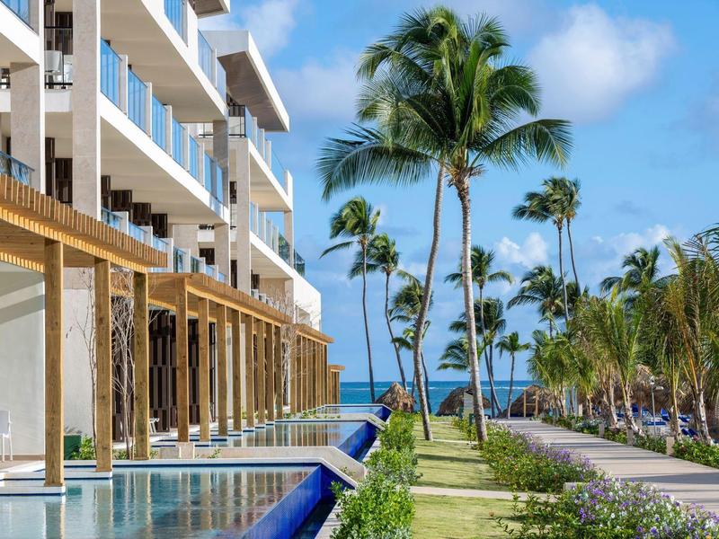 Modernes Hotel mit Pool, Palmen und blauem Himmel entlang eines grünen Gartens am Meer.
