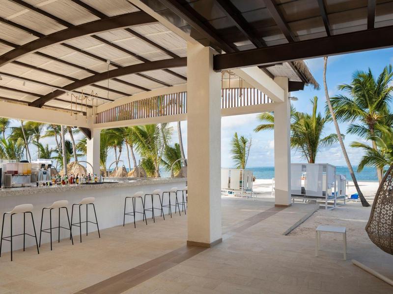 Geöffnete Strandbar mit Hockern, Palmen im Hintergrund und Blick auf das blaue Meer.