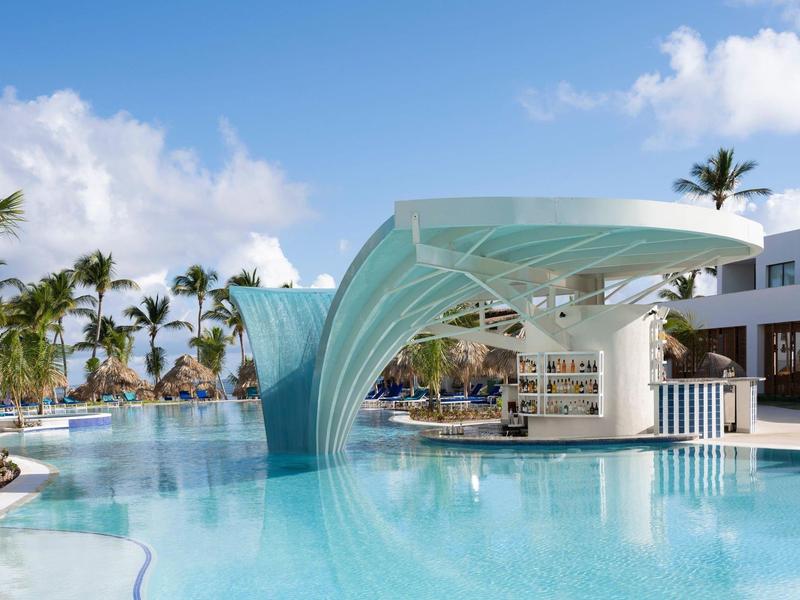Moderner Pool mit Wasserfallbar, Palmen und Liegestühlen unter blauem Himmel.