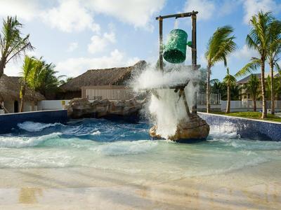 Wasserbecken mit schwebendem grünen Eimer, der Wasser über eine Felsstruktur schüttet, Palmen im Hintergrund.