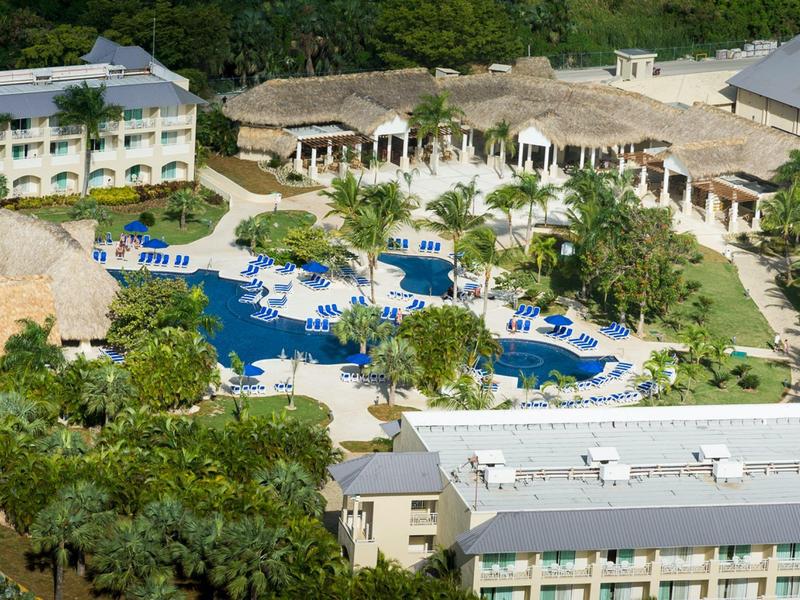 Hotelanlage mit mehreren Gebäuden, großem Pool, blauen Liegestühlen und grünen Bäumen.