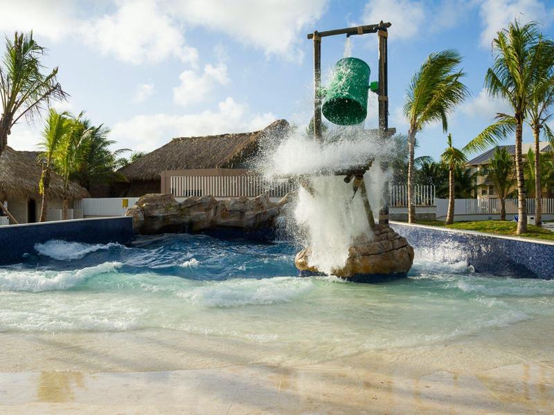 Wasserbecken mit schwebendem grünen Eimer, der Wasser über eine Felsstruktur schüttet, Palmen im Hintergrund.