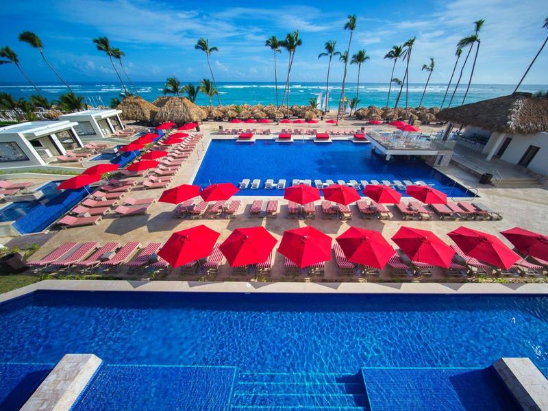 Velká oblast bazénu s červenými slunečníky, lehátky a palmami za slunečného dne.