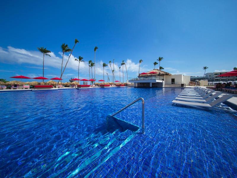 Velký modrý bazén s červenými slunečníky a palmami pod jasnou oblohou.