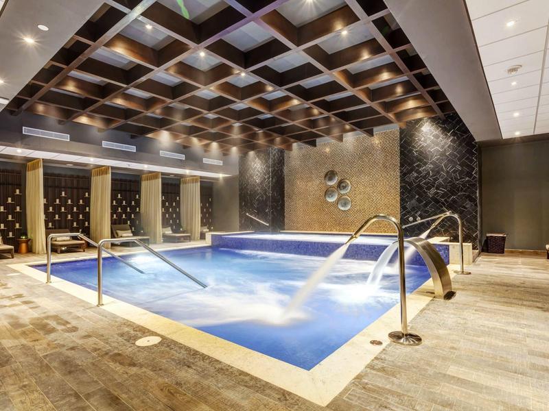 Moderní vnitřní bazén s vodními fontánami, dřevěným stropem a pohodlnými posezeními v hotelu.