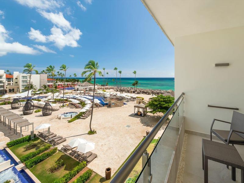 Balkon se dvěma židlemi s výhledem na hotelový bazén, lehátka, pláž a moře pod modrou oblohou.