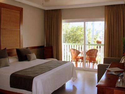 Modern hotel room with large double bed, balcony, and greenery view.