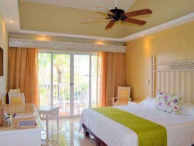 Bright hotel room with double bed, chairs, ceiling fan, and balcony overlooking garden.