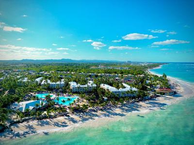 Aerial view of a beach resort with pools, palm trees, and clear sea under a blue sky.
