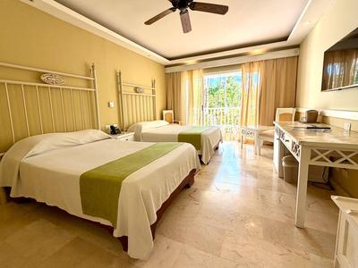 Bright hotel room with two beds, ceiling fan, and balcony overlooking green areas.