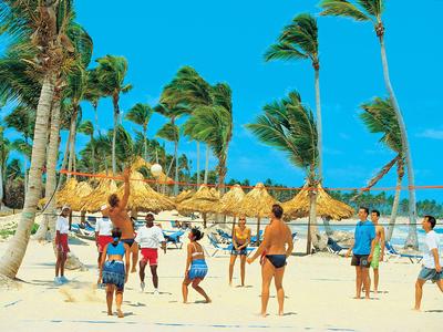 People playing beach volleyball on sandy shore with palm trees and sun umbrellas.
