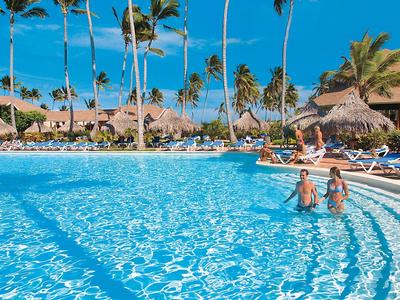 Large pool with crystal-clear water, surrounded by lounge chairs and palm trees under blue sky.