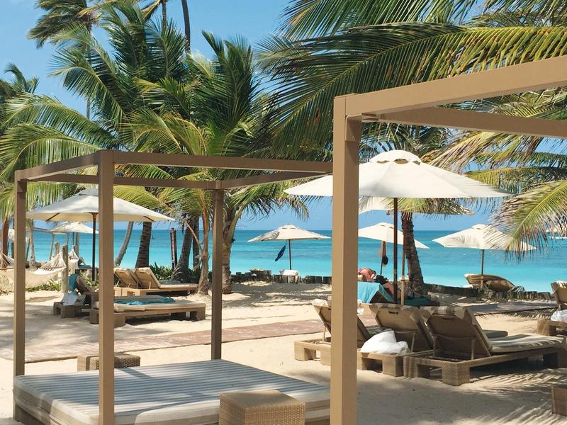 Beach with wooden cabanas, loungers, and white umbrellas under palm trees by the sea.