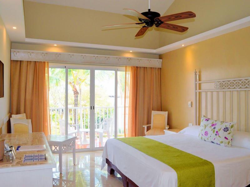 Bright hotel room with double bed, chairs, ceiling fan, and balcony overlooking garden.