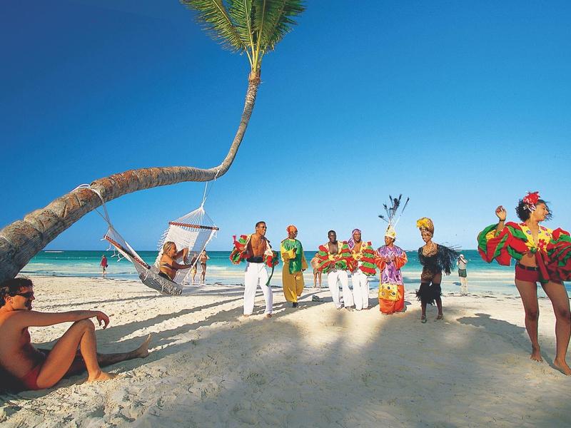 Beach with white sand, palm trees, and a group of musicians and dancers in colorful costumes.