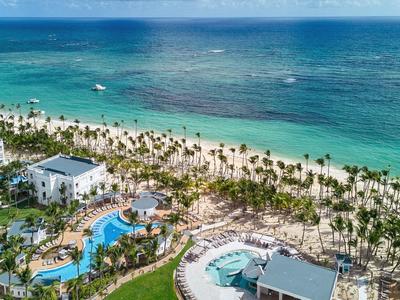 Aerial view of a resort with pools, palm trees, and a long sandy beach by the turquoise sea.