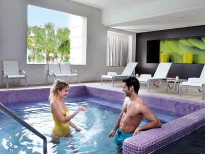 Two people in an indoor hotel pool with lounge chairs and green plants in the background.