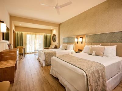 Bright hotel room with two double beds, wooden floor, and balcony access.