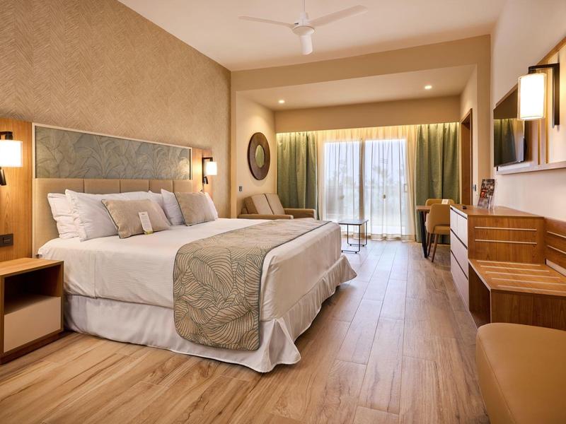 Bright hotel room with large bed, wooden floor, and balcony overlooking the garden.