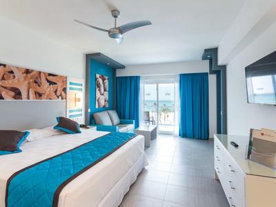 Bright hotel room with double bed, blue curtains, and balcony overlooking the sea.