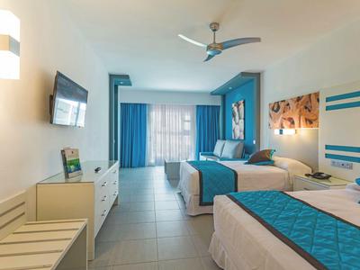 Jasny pokój hotelowy z dwoma łóżkami, niebieskimi zasłonami i nowoczesnym wyposażeniem.