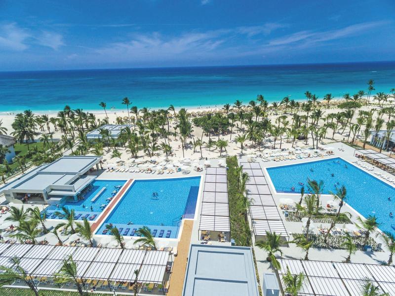 View of hotel pools, palm trees, and a sandy beach with blue sea in the background.