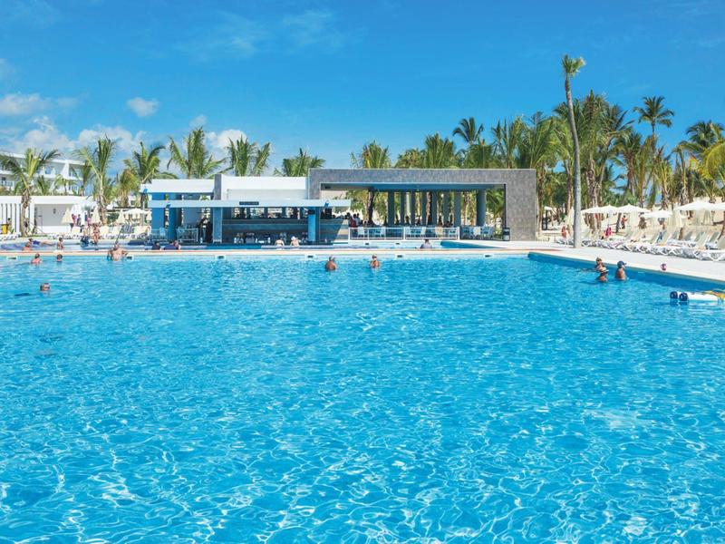 Large hotel pool with clear blue water, surrounded by white lounge chairs and palm trees.