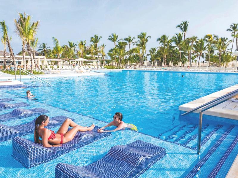 Large hotel pool with loungers in water and palm trees in the background on a sunny day.