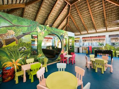 Světlé dětské hřiště s barevnými stoly a židlemi v tropickém hotelovém interiéru.