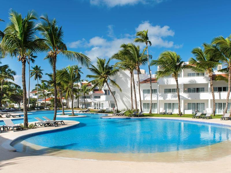 Velký hotelový bazén s palmami a lehátky pod modrou oblohou.