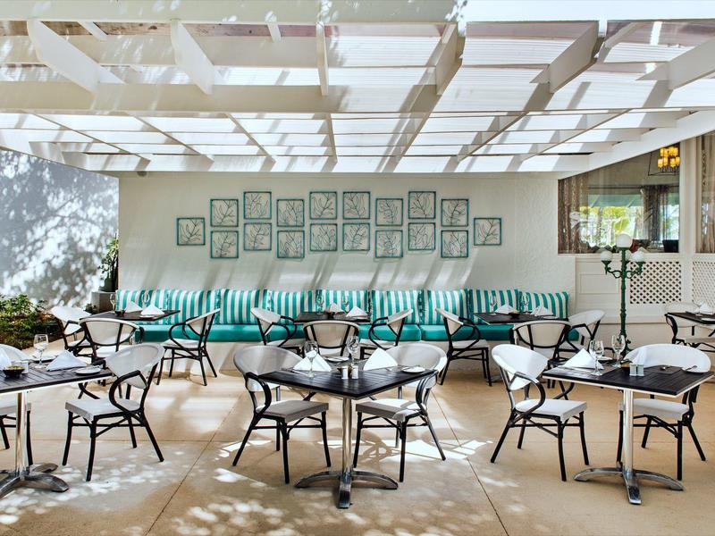 Světlá terasa s černými židlemi, kulatými stolky a dlouhou lavicí s tyrkysovými polštáři.