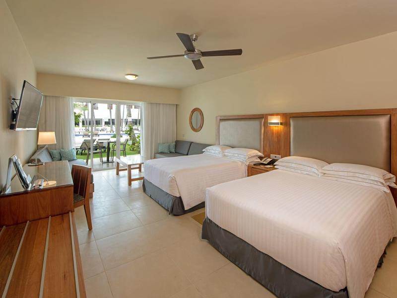 Světelný hotelový pokoj se dvěma velkými postelemi, stropním ventilátorem a výhledem na balkon.