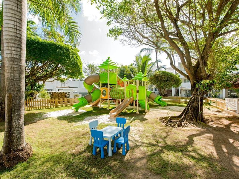 Slunné hřiště s lezeckou konstrukcí a dvěma malými stolky se židlemi pod velkými stromy.