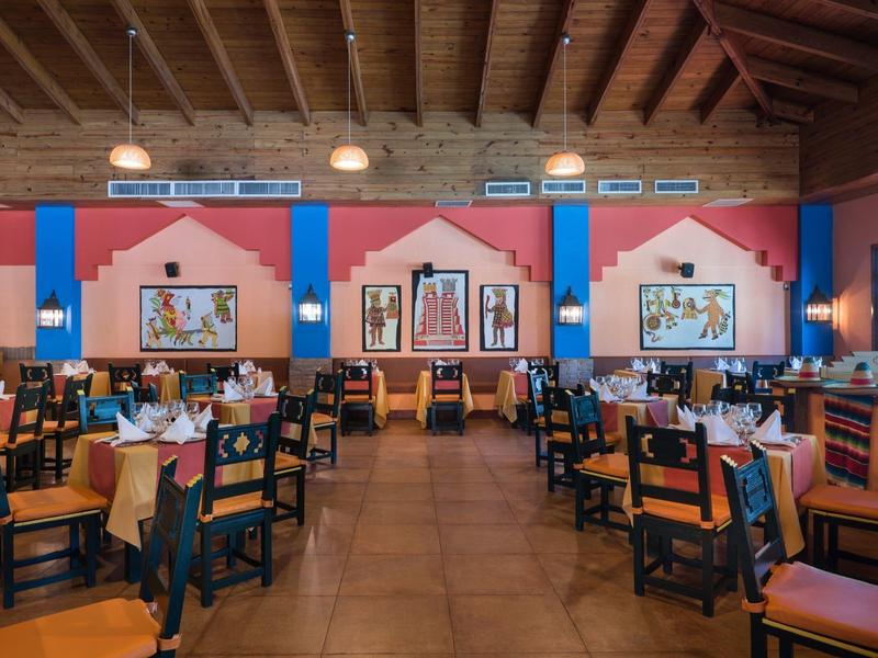 Velká restaurace s dřevěným stropem, barevnými zdmi a mnoha stoly a židlemi.