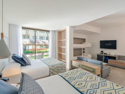 Bright hotel room with double beds, bunk bed, TV, and garden and pool view.