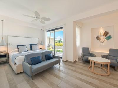 Bright hotel room with double bed, sofa, two chairs, and small round table near balcony