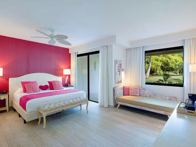 Bright hotel room with double bed, pink wall, window with green view, bench and desk.