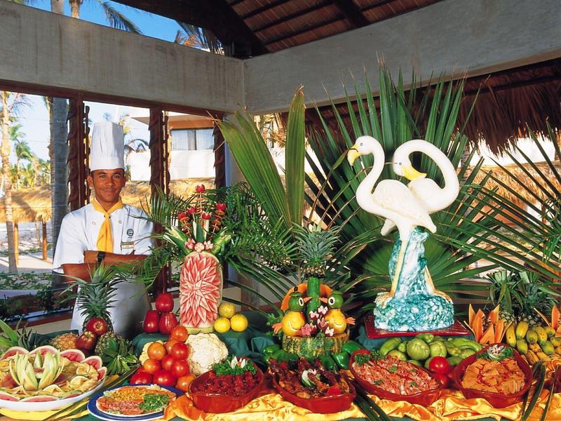 Buffet with tropical fruits and artistic sculptures, chef in the background under a wooden roof.