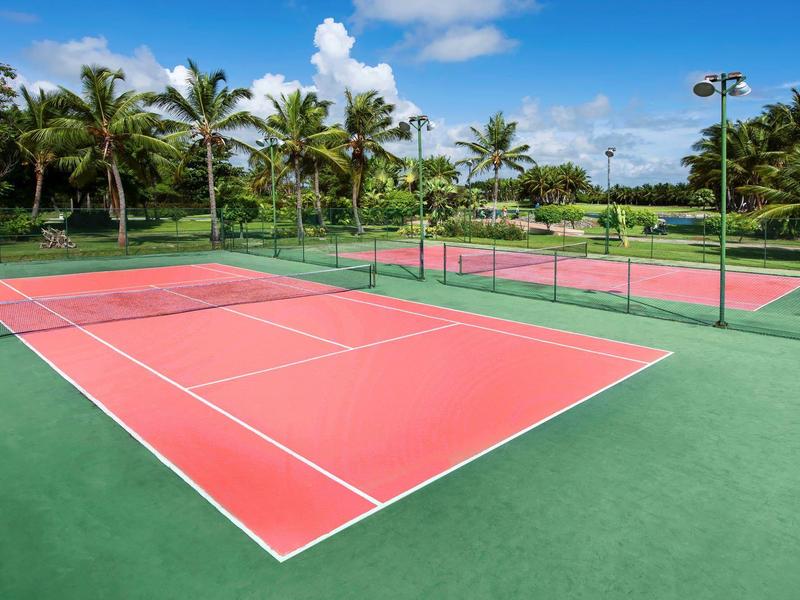 Red tennis courts with green borders surrounded by palm trees under a blue sky.